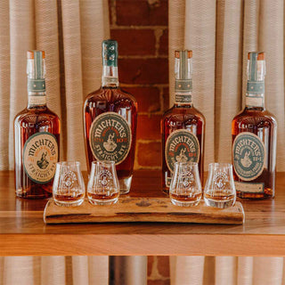Four bottles of Michter’s US*1 Kentucky Straight Rye Whiskey rest on a wooden shelf, five glasses before them on a tray—each offering notes of butterscotch and vanilla. Beige curtains and a brick wall set the inviting scene.