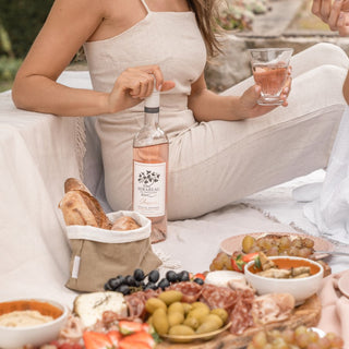 Woman enjoying a picnic with a bottle of Mirabeau Rosé and assorted foods on a blanket outdoors.