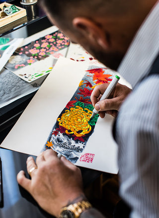 A close-up lifestyle shot of an artist drawing a detailed, colorful illustration of a chrysanthemum and dragon on white paper using a green-tipped marker.