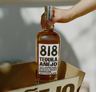 A hand lifts a 700ml bottle of 818 Tequila Anejo from a cardboard box, its label with bold black text and details on a white background, hinting at notes of honeyed agave and toasted oak.