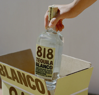 A hand lifts a clear bottle of 818 Tequila Blanco 700ml with a beige label from an open box marked BLANCO, in a brightly lit, minimalistic scene that highlights this smooth tequila's fresh agave and citrus notes.
