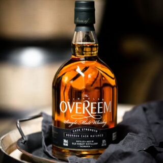 A bottle of Overeem Bourbon Cask – Cask Strength Tasmanian Single Malt Whisky rests on a metal tray beside a dark cloth. Its amber liquid, in a clear glass bottle with black and gold label and cap, suggests notes of vanilla, citrus, and malt oak.