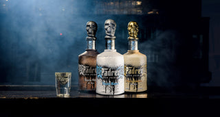 Three Pādre Azul Super Premium Tequila Reposado 700ml bottles, each with a metallic skull cap in brown, white, and cream, stand on a dark bar beside a branded shot glass amid mist and hints of vanilla caramel and cooked agave.
