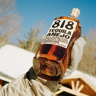A gloved hand holds a bottle of 818 Tequila Anejo 700ml outdoors, snowy roof and trees behind, beneath a clear blue sky—ideal for sipping and enjoying its honeyed agave and toasted oak notes.