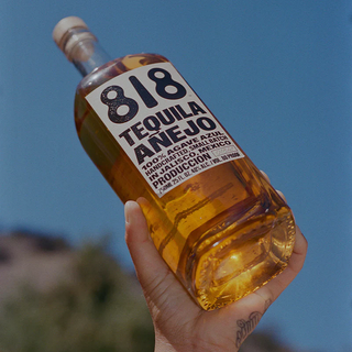 A hand holds a bottle of 818 Tequila Anejo 700ml against a clear blue sky, its amber liquid shining. Made from 100% agave azul and handcrafted in Jalisco, Mexico, the label reveals notes of honeyed agave and toasted oak.