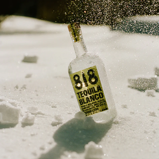A bottle of 818 Tequila Blanco 700ml stands partly buried upright in the snow, while sunlight highlights snowflakes swirling in the air around it.