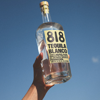 A hand holds up a clear bottle of 818 Tequila Blanco 700ml against a vivid blue sky. Its light yellow label with bold black text showcases the tequila’s fresh agave notes and smooth, crisp profile.