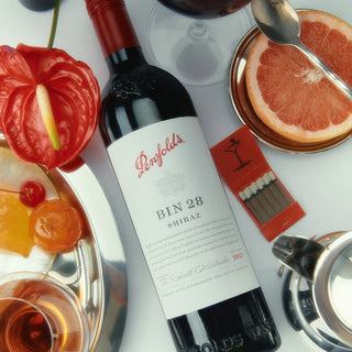 Penfolds Bin 28 Shiraz wine bottle with fruit and a glass on a white surface