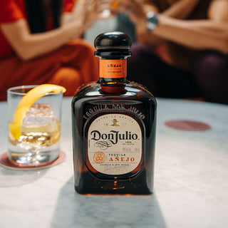 A bottle of Don Julio Anejo Tequila 700ml rests on a marble table beside a whiskey glass garnished with lemon peel, while two people chat in the softly blurred background.