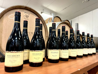 A row of Rockburn Central Otago Pinot Noir wine bottles displayed on a wooden table in front of large wooden wine barrels.