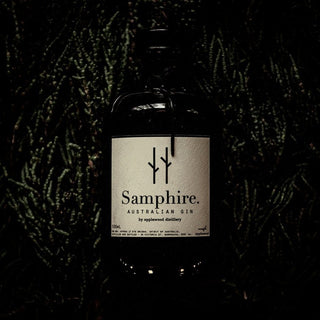 A dark glass bottle of Applewood Samphire Gin with a white label, minimal black text, and design elements sits against leafy greenery, hinting at its coastal botanicals and fresh lemon myrtle inside.