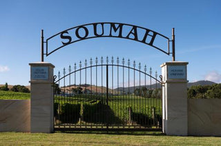 A large black metal arched gate with the word 'SOUMAH' across the top, set between two beige stone pillars. Beyond the gate is a vineyard landscape with green grapevines, hills, and a blue sky.