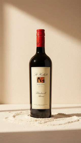 A bottle of St Malickt Barolo Blackfell Sherry red wine stands on a sandy surface against a warm beige wall with dramatic diagonal shadows.
