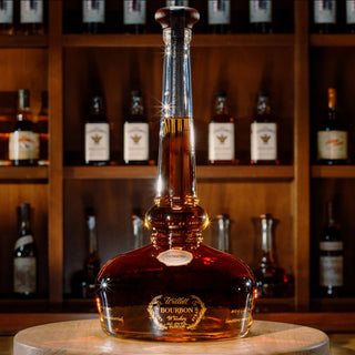 A bottle of Willett Family Pot Still Reserve Bourbon Whiskey rests on a wooden table, its tall, elegant neck and gold accents suggesting rich notes of vanilla and toasted oak, with shelves of assorted liquor bottles in the background.