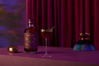A bottle of Four Pillars Vodka and Vinegar cocktail mix, shown next to a filled coupe glass with a green garnish, set on a purple table against a dark red curtain background.